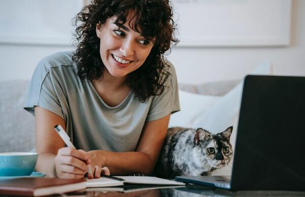 A imagem contém uma estudante sorrindo e olhando para o notebook. Ela faz anotações em companhia de seu gato de estimação.