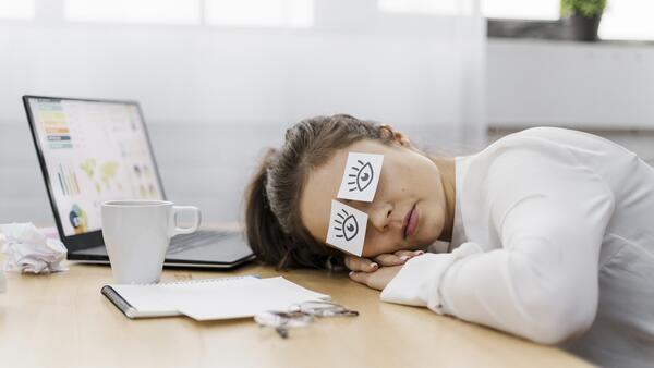 A imagem contém uma moça com a cabeça deitada sobre os braços em sua área de trabalho. Em seus olhos fechados estão grudados adesivos de olhos abertos, indicando Síndrome do Impostor.