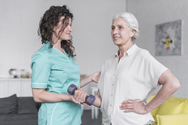 A imagem contém uma senhora fazendo fisioterapia geriátrica. Sua terapeuta dá apoio para ela enquanto ela levanta um pesinho de mão.