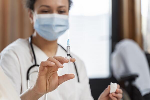 A imagem contém uma enfermeira, em desfoque, com uma seringa na mão apontada para cima. A enfermeira aguarda os próximos pacientes segundo a tabela de vacinação.