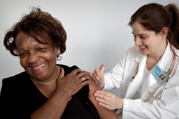 A imagem contém uma senhora tomando vacina após sua idade ser liberada de acordo com a tabela de vacinação.