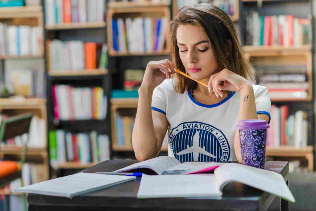 Aluna estudando na biblioteca, olhando para o livro aberto sob a mesa. 