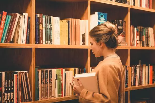 A imagem contém uma estudante escolhendo livros em uma estante de biblioteca.