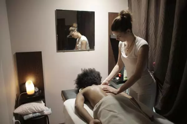 A imagem apresenta uma sala de massoterapia. Na esteira uma pessoa está deitada de costas, enquanto a fisioterapeuta dermatofuncional massageia suas costas.